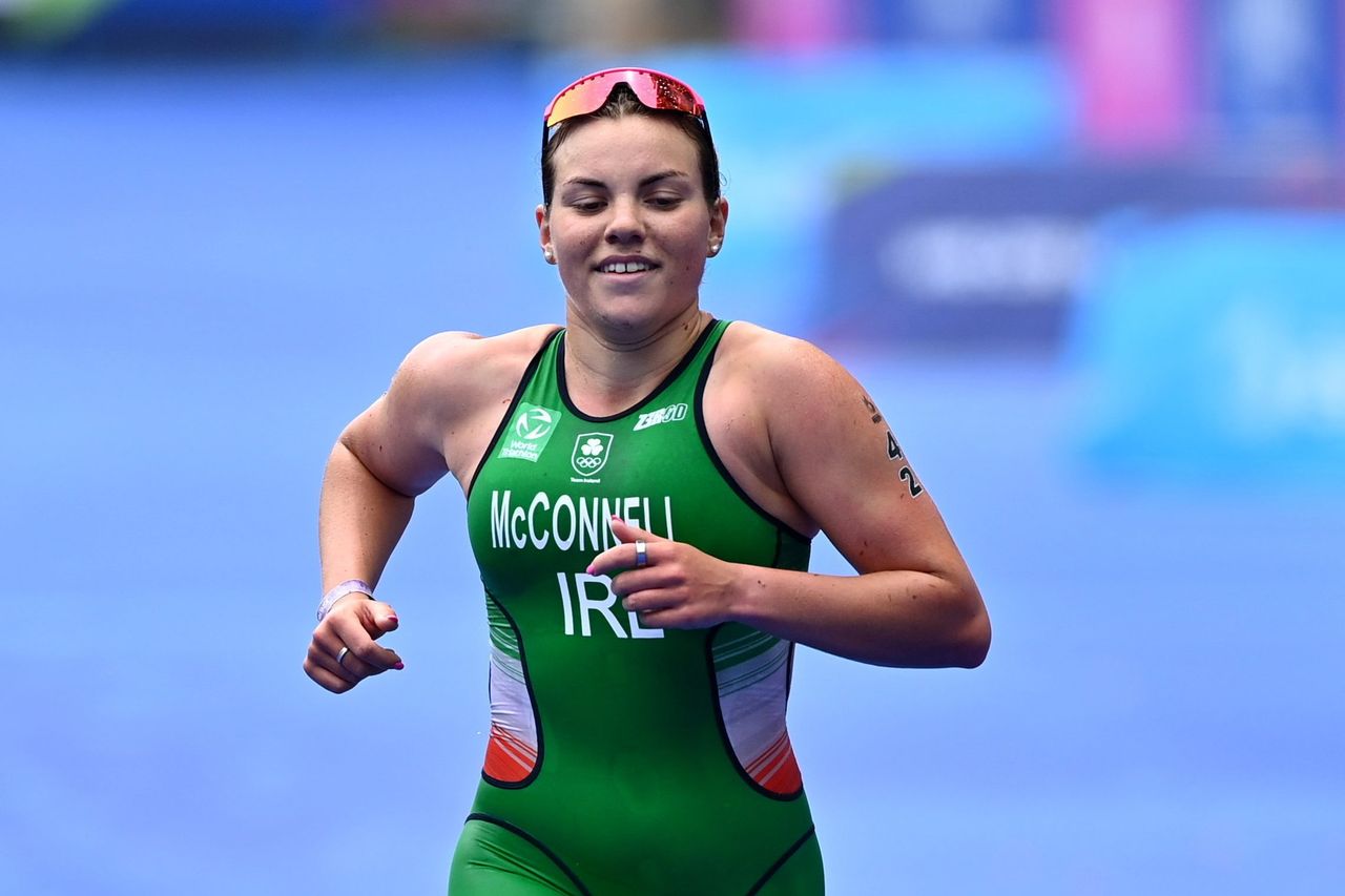 Erin McConnell, Irish triathlete, competing in a race. She wears a green Ireland triathlon suit with her name printed on the front, running with determination. The background features a blue racecourse, highlighting the competitive atmosphere.