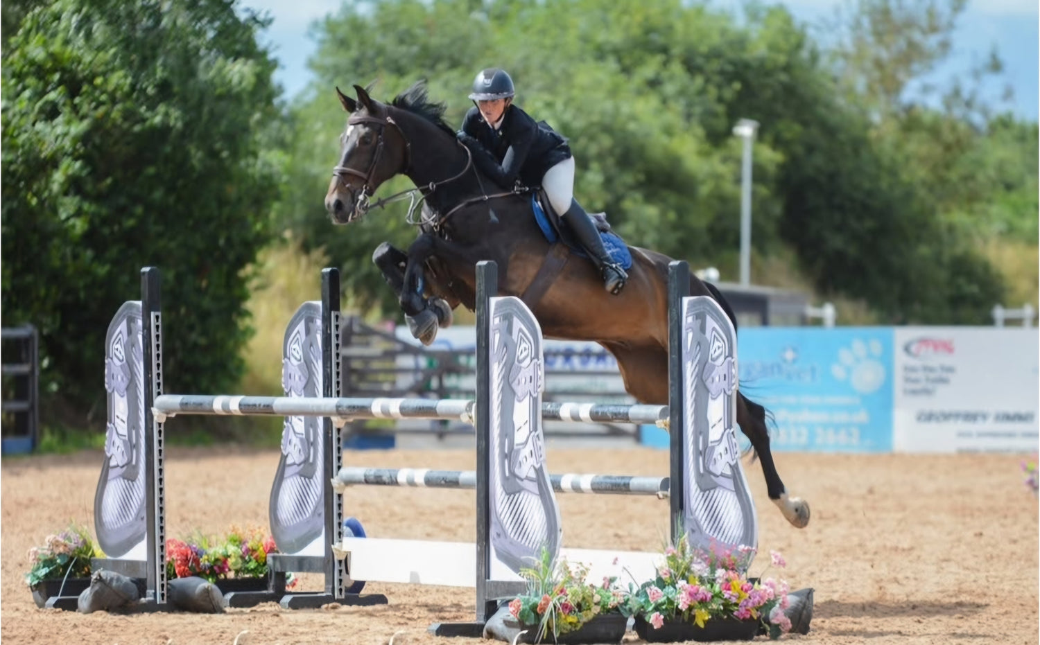 Sonya McAleer mid-air during a showjumping competition, riding a powerful dark bay horse over a decorated jump. Dressed in traditional equestrian attire with a helmet and navy saddle pad, she demonstrates focus and precision in the arena.
