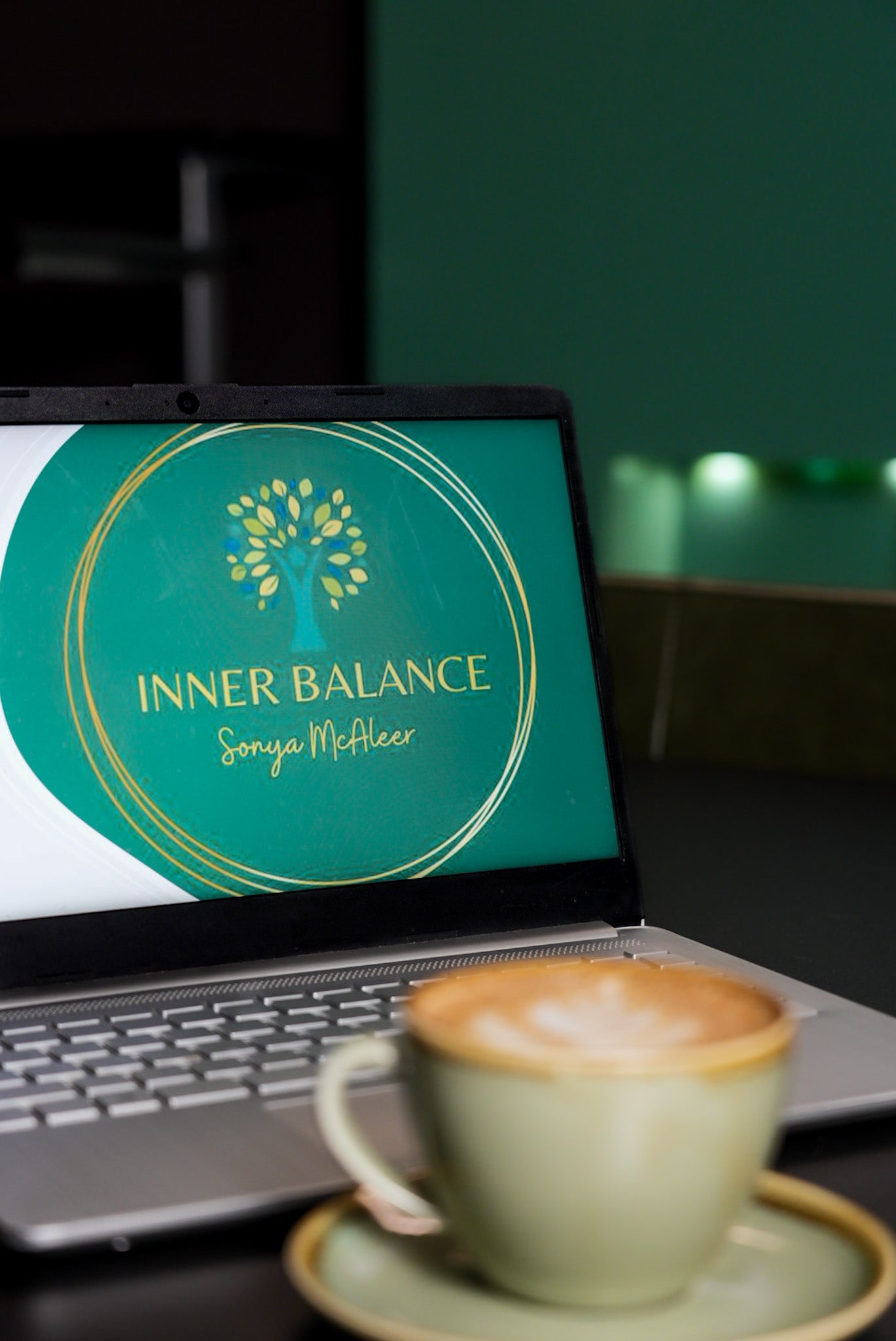A laptop displaying the "Inner Balance - Sonya McAleer" logo on a green background with gold accents. In the foreground, a beautifully crafted latte in a matching green cup sits on a saucer, creating a calm and focused workspace ambiance.