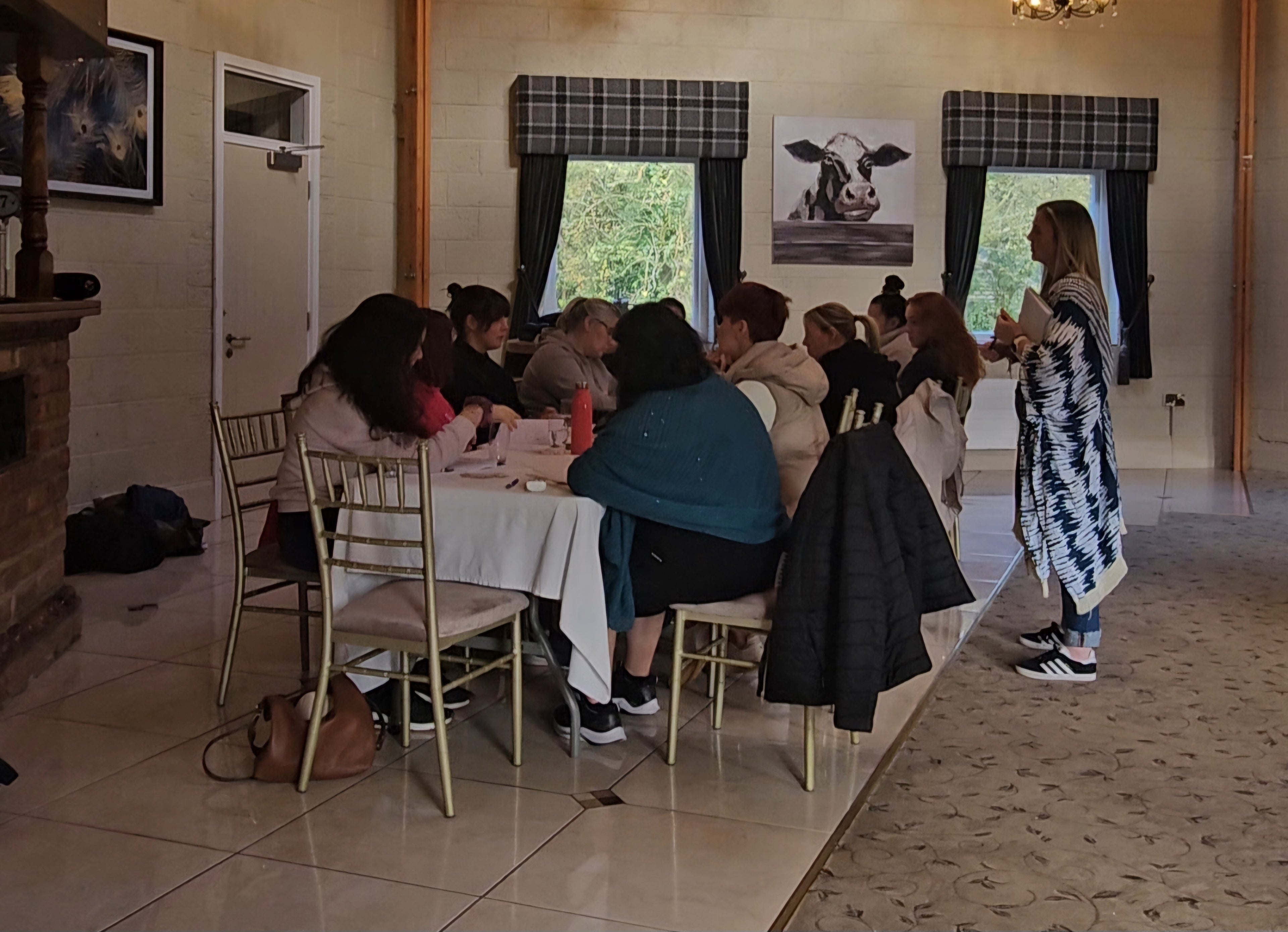 Sonya McAleer leading a group workshop in a cozy retreat setting. Participants sit around a table, engaged in discussion and reflection, while Sonya stands, guiding the session with a notebook in hand. The room features warm lighting, rustic decor, and a peaceful atmosphere