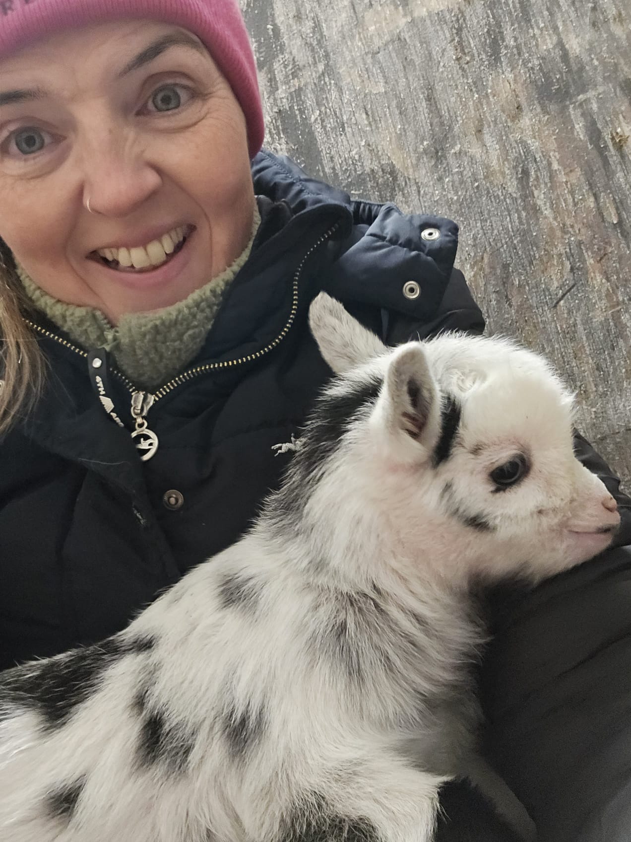 Sonya McAleer Smiling while holding an adorable black-and-white baby goat. Dressed in warm outdoor clothing, she embodies the connection between women in farming and agriculture.