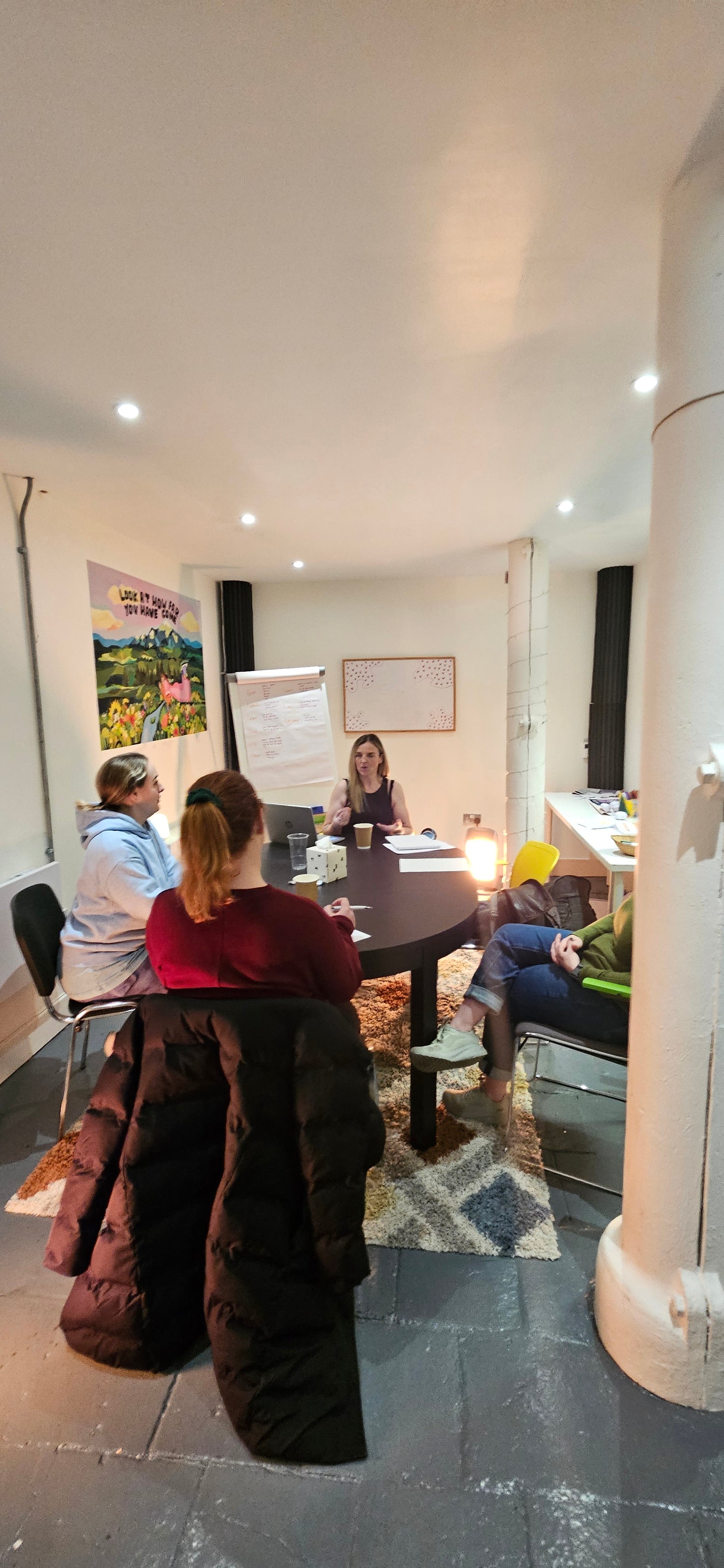 Sonya McAleer leading an intimate coaching session in a cozy, creative space. She sits at a round table, engaging with a small group of women, fostering deep conversation and personal growth. The warm lighting and relaxed atmosphere create a supportive and inspiring environment.