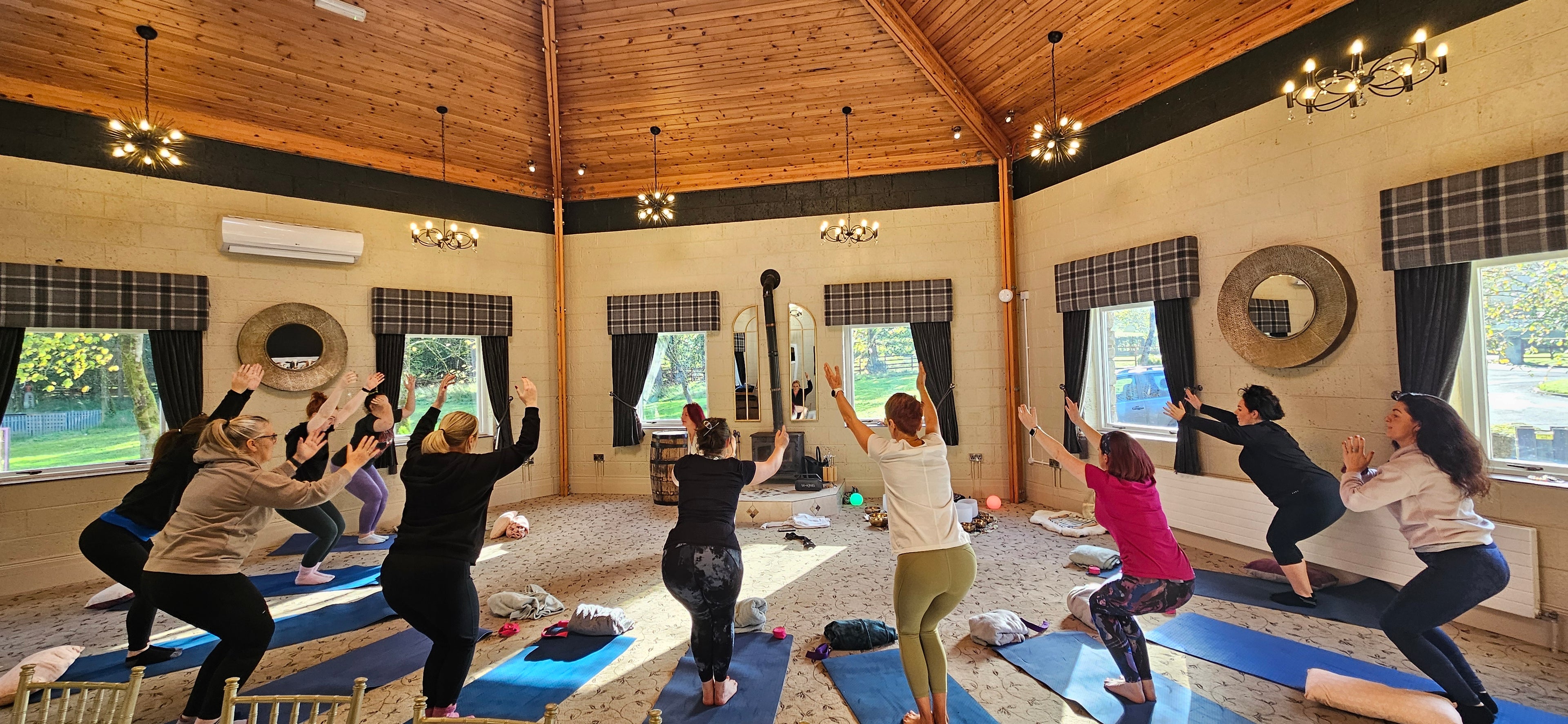 A group of women participating in a yoga session inside a beautifully lit retreat space with wooden ceilings and large windows overlooking nature. They are on yoga mats, practicing a pose with arms raised, embracing movement, mindfulness, and connection in a peaceful environment.