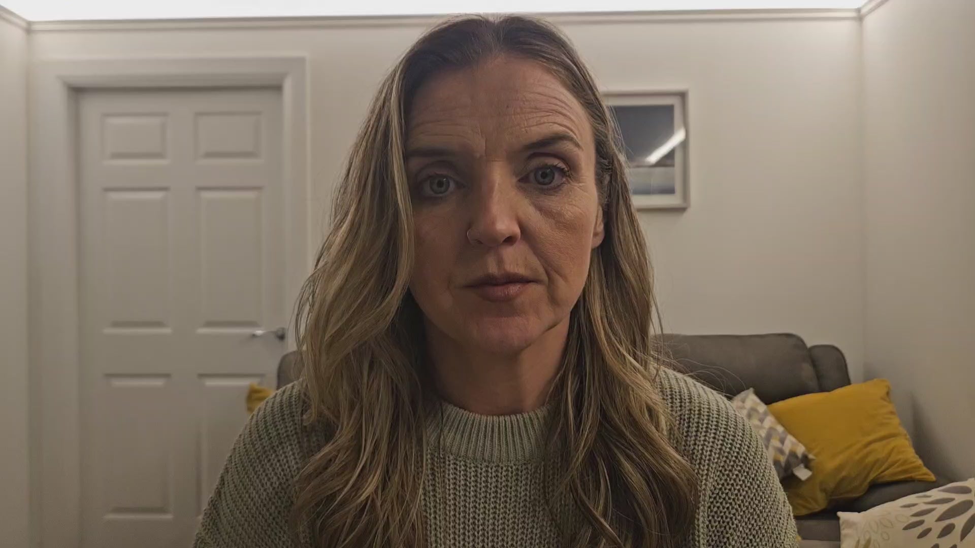 Load video: Sonya McAleer sitting in a cosy setting, smiling warmly. She wears a light green jumper.. The background features a white wall, grey sofa with yellow cushions creating a relaxed and inviting atmosphere.