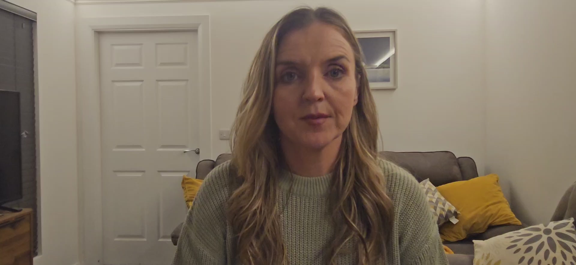 Load video: Sonya McAleer sitting in a cosy setting, smiling warmly. She wears a light green jumper.. The background features a white wall, grey sofa with yellow cushions creating a relaxed and inviting atmosphere.